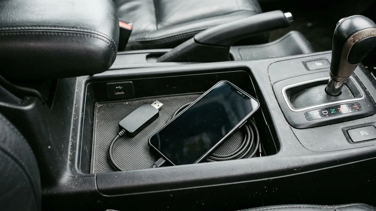Parked car center console cubby showing an unplugged matte-black wireless USB adapter, a coiled braided USB-C cable, and a dark-screen smartphone on a rubber tray