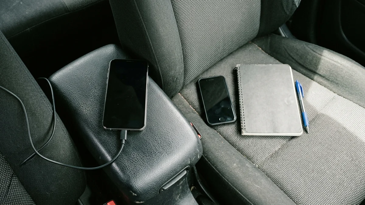 A cabled dark-screen phone on a car armrest and a second phone beside a notebook on the passenger seat.